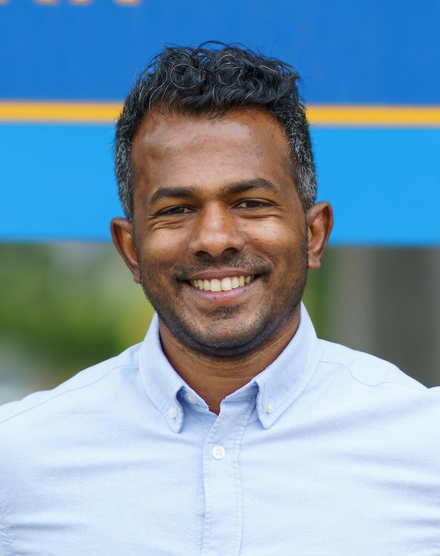 Sujan is wearing a blue button shirt, he has salt and pepper hair and is smiling.